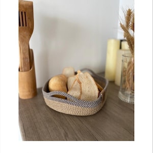 May include: A square jute and cotton bread basket with a gray and white pattern. The basket is filled with bread and is sitting on a wooden table.