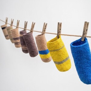 May include: A series of cylindrical crochet pouches in various colors including blue, yellow, tan, and brown, each hanging from a horizontal twine string by a wooden clothespin. The pouches feature different striped patterns and solid textures.