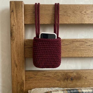 May include: A burgundy crochet phone holder with matching braided straps, hanging from a wooden headboard. The holder contains a white and black smartphone. The bed has a plaid pillow and blanket.