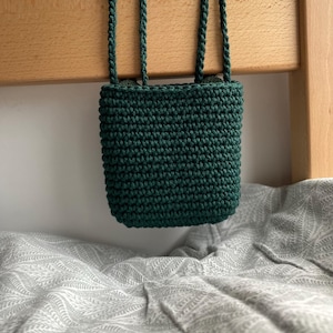 May include: A small, dark green crochet bag with four braided handles. The bag is rectangular and hangs from a wooden beam. The background features a patterned, gray and white fabric.