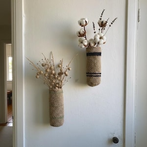 Op de afbeelding: Twee cilindervormige wandplantenbakken gemaakt van geweven beige juteweefsel. De linker plantenbak heeft een subtiele groene horizontale streep, terwijl de rechter twee duidelijke zwarte horizontale strepen heeft. Beide bevatten droogbloemen.