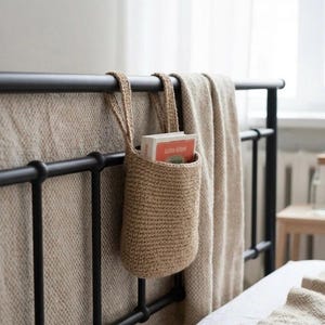 May include: A woven, beige bedside organizer hanging from a black metal bed frame. The organizer holds several books with orange and white covers. A beige blanket is draped over the bed frame, and the bed has white linens.