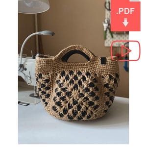 May include: A jute crochet tote bag with a black floral pattern. The bag has a rounded shape and black handles. The text "DIY JUTE CROCHET TOTE BAG" is at the bottom. The bag is on a white surface.
