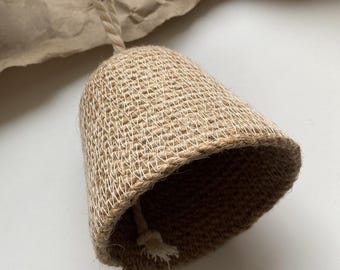 May include: A woven jute bell with a natural fiber rope for hanging. The bell has a flared shape and a small loop at the bottom. The background includes a sprig of greenery and a piece of brown paper.