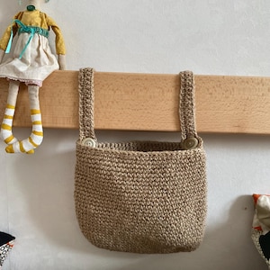 Könnte beinhalten: Eine kleine, gewebte Jutetasche mit zwei Knopfverschlüssen und zwei gewebten Trägern. Die Tasche hat eine natürliche, beige Farbe und hängt an einem Holzbalken. Eine handgefertigte Puppe mit gelbem Oberteil und gestreiften Beinen sitzt auf dem Balken.