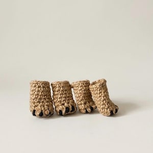 Puede incluir: Cuatro pequeños botines de ganchillo en color tostado, diseñados para parecer patas de animal. Cada botín presenta una construcción de hilo tejido con textura y detalles distintivos en hilo negro en la base para representar los dedos.