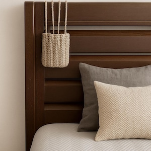 Puede incluir: Una cesta beige tejida con un asa de cuerda cuelga de un cabecero de madera marrón oscuro. La cama tiene un edredón blanco estampado y dos almohadas, una gris y otra crema con un diseño de espiga. La escena evoca calma.