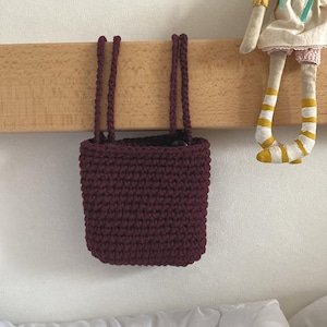 Può includere: Una borsa all'uncinetto bordeaux con manici coordinati è appesa a una trave di legno. La borsa è di forma rettangolare con il fondo arrotondato. Sullo sfondo è visibile una bambola fatta a mano con un top giallo e calzini a righe.