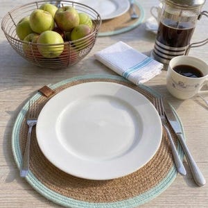 Op de afbeelding: Een tafelsetting met een wit bord, bestek en een geweven placemat met een lichtblauwe rand. Een draadmand bevat groene en rode appels. Een cafetière en een kopje koffie staan ook op de tafel.