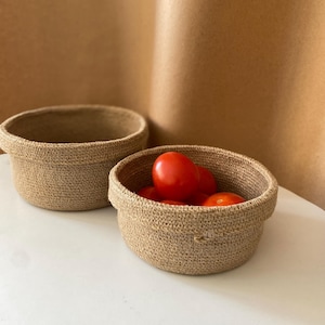 Op de afbeelding: Twee geweven jute manden. Eén mand is leeg en de andere bevat verschillende rode tomaten. De manden zijn rond met kleine handvatten en staan op een wit oppervlak tegen een bruine achtergrond.