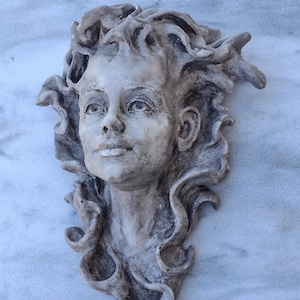 Pode incluir: Uma escultura de gesso branco de um rosto de mulher com cabelo solto. A escultura está montada em uma parede e tem uma aparência realista.