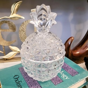 May include: A clear glass pineapple-shaped container with a textured surface. The container is sitting on top of a teal book with the word "Oxford" printed on the cover.