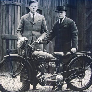 May include: Vintage black and silver motorcycle with two men in suits. The motorcycle has a round gas tank, a single seat, and spoked wheels. The men are standing next to the motorcycle, posing for a photograph.