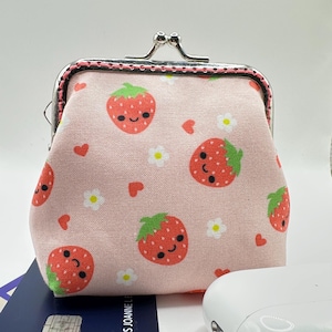 Puede incluir: Un monedero rosa con cierre plateado, con un estampado repetido de fresas sonrientes, flores blancas y corazones rojos. El bolso tiene un cierre de beso y un ribete rosa y negro.