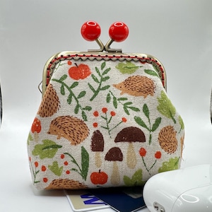 May include: A whimsical coin purse with a red clasp and a fabric design featuring hedgehogs, mushrooms, and foliage in orange, green, and brown. The purse has a metal frame and red stitching.