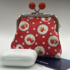 May include: A red coin purse with a strawberry pattern, a white case, and a blue card. The purse has a silver frame and red ball closures. The strawberry pattern is on a white background.