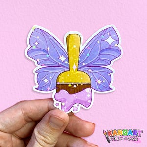 May include: A purple and gold paintbrush with glitter and a butterfly design. The paintbrush is dripping with pink paint and has a glittery yellow handle.