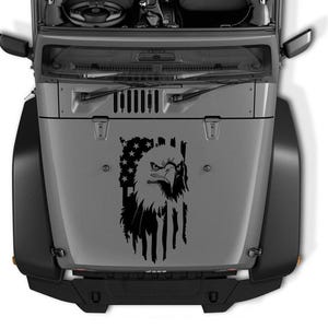Puede incluir: Calcomanía en blanco y negro de un águila calva americana con la cabeza girada hacia un lado, posada sobre una bandera estadounidense desgarrada. La calcomanía se aplica al capó de un Jeep Wrangler gris.