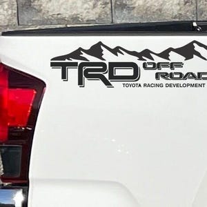 Op de afbeelding: Zwarte TRD Off Road sticker met een bergketenafbeelding, aangebracht op de witte zijkant van een vrachtwagen. De tekst "TOYOTA RACING DEVELOPMENT" staat eronder.