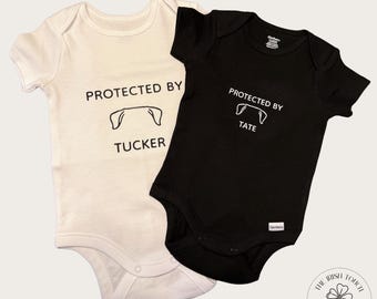 May include: Two white baby onesies with black text. One onesie says "Protected by Tucker" with a dog paw print. The other onesie says "Protected by Tate" with a dog paw print.