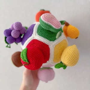 Könnte beinhalten: Ein mehrfarbiger Häkelball mit verschiedenen dreidimensionalen Fruchtformen auf der Oberfläche, darunter violette Weintrauben, ein roter Apfel, eine gelbe Zitrone, eine Orange, eine rosa Birne und eine braune Kiwi, verbunden durch weiße Nähte.