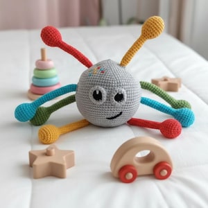 Puede incluir: Un juguete de bebé de ganchillo gris con apéndices coloridos y una cara sonriente. El juguete tiene brazos rojos, amarillos, verdes y azules. También hay juguetes de madera, como una estrella, un coche y un anillo apilable.