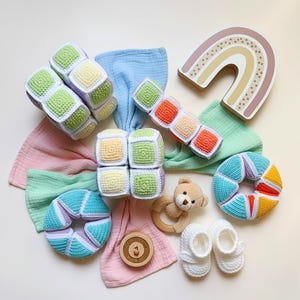Puede incluir: Colección de juguetes para bebés de colores pastel, que incluye bloques de ganchillo, un juguete de madera con forma de arcoíris, un mordedor de madera y un par de zapatos de bebé de ganchillo blancos. También se incluyen muselinas suaves.