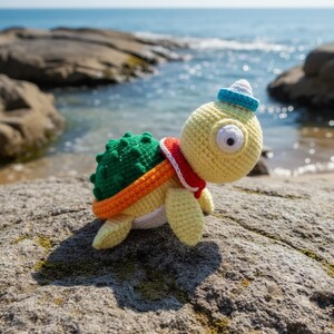 May include: A handmade crochet turtle toy with a yellow body, green shell, and orange and red accents. The turtle wears a blue and white hat and a red scarf. The toy is set on a rock near the ocean.