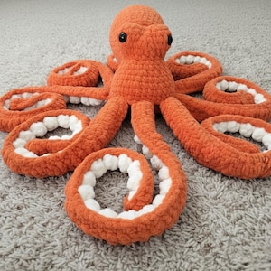May include: A plush octopus toy crafted from thick, orange yarn with a soft texture. The creature features a rounded head with two black, circular eyes and eight curled tentacles. Each tentacle displays a contrasting white, textured underside.