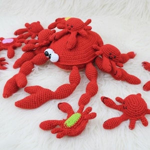 May include: A group of handmade red crochet crab toys with white eyes and various accent colours. The crabs vary in size, with a large central crab surrounded by smaller ones. The toys are displayed on a white, fluffy surface.
