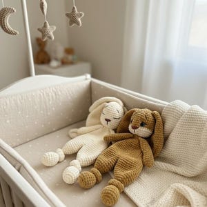 Puede incluir: Dos peluches de conejo tejidos a ganchillo, uno en color crema y otro en marrón, con orejas largas y caídas, rasgos faciales bordados y cuerpos de textura suave. Estos juguetes están colocados en una cuna blanca con forro beige.