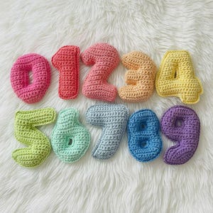 May include: A set of ten crochet numbers from zero to nine, each crafted in a different solid color including pink, red, coral, peach, yellow, lime green, mint, light blue, medium blue, and purple, featuring a textured, hand-stitched yarn finish.