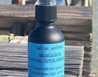 Op de afbeelding: Een zwarte fles met een doorzichtige spuitmond, met het label "Salt air, wellness Magnesium Oil All Natural Topical Mineral". De fles staat op een houten oppervlak.