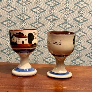 May include: Two vintage ceramic egg cups. One cup features a painted house and the text "Kent Cafe". The other cup has the words "Just Laid". Both cups have a white base with blue and brown accents.