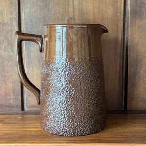 May include: A brown ceramic pitcher with a textured lower half and a smooth upper half. The pitcher has a handle and a spout for pouring. The pitcher is sitting on a wooden shelf.