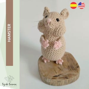 Puede incluir: Un juguete de hámster de crochet con un cuerpo beige y patas rosas, de pie sobre una base de madera. El juguete está hecho a mano y tiene una expresión linda y amigable.