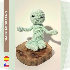 May include: A crocheted green figure with closed eyes meditating in a seated position on a wooden log. The figure has a simple smile.