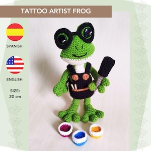 May include: Green crocheted frog wearing glasses and a black and orange jumpsuit, holding a tattoo needle. The frog is surrounded by small bowls of paint in red, blue, and yellow.