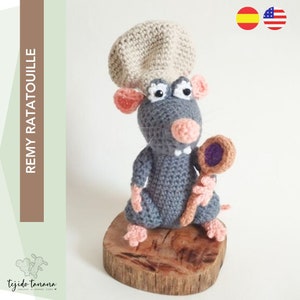 May include: A crocheted rat wearing a chef's hat and holding a wooden spoon. The rat is gray with pink ears and feet. It is sitting on a wooden base.