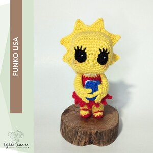 Puede incluir: Una figura de Lisa Simpson Funko Pop de crochet amarillo sosteniendo una pelota azul y roja. La figura está de pie sobre una base de madera.