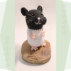 May include: A crocheted black and white hamster wearing a white crocheted shirt. The hamster is standing on a wooden base.
