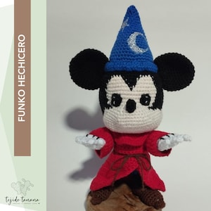 Puede incluir: Un Mickey Mouse de crochet vestido de mago. Lleva un sombrero azul con una estrella y una luna creciente, una túnica roja y zapatos marrones. Está de pie sobre una base de madera.