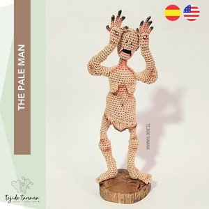May include: A crocheted figure of a pale man with large eyes and a wide mouth, standing on a wooden base. The figure is beige and has a textured surface. The text "THE PALE MAN" is written vertically on the left side of the image.