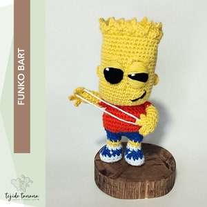 Puede incluir: Una figura Funko Pop de Bart Simpson de Los Simpson hecha a crochet. Lleva una camisa roja, pantalones cortos azules y zapatillas blancas. Está sosteniendo una honda en su mano derecha. La figura está de pie sobre una base de madera.