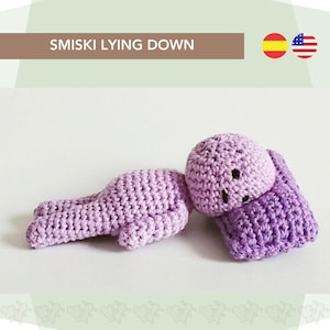 May include: A purple crocheted amigurumi doll lying down on a purple crocheted pillow. The doll has a simple face with two black eyes and a small mouth.