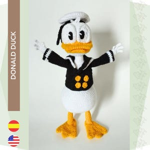 以下が含まれることがあります： 白い帽子、黒いジャケット、黄色のボタンが付いたセーラー服を着た、かぎ針編みのドナルドダック人形。人形は白い体、オレンジ色のくちばし、黄色の足を持っています。