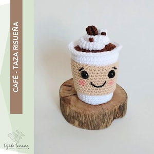 Puede incluir: Una taza de café de ganchillo con una cara sonriente, crema batida y granos de café en la parte superior. La taza está sentada sobre una rodaja de madera.