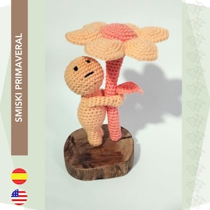 Peut inclure: Une poupée amigurumi en crochet avec un corps de couleur pêche et une fleur sur la tête. La poupée tient la tige de la fleur avec ses bras. La poupée est debout sur une base en bois.