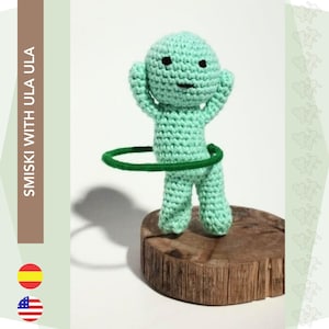 May include: A green crocheted amigurumi doll with a green hula hoop around its waist. The doll is standing on a wooden base.