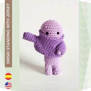 Puede incluir: Un juguete de amigurumi de color morado tejido a crochet con un suéter y una cara simple. El juguete está de pie sobre una superficie blanca.